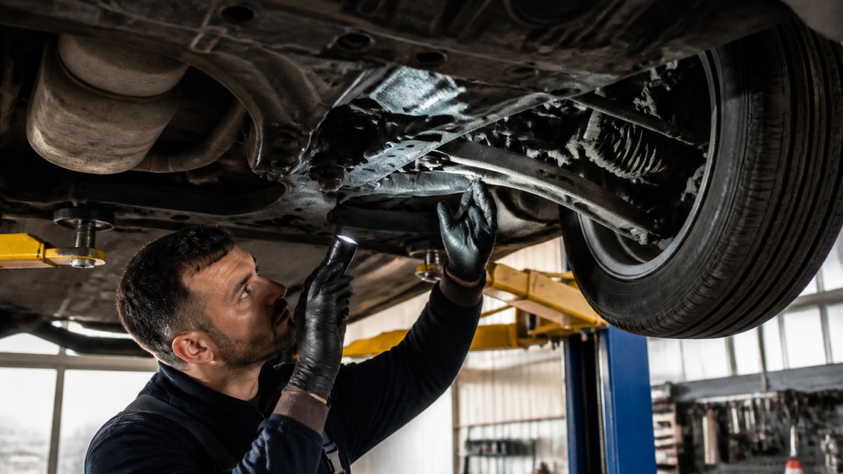 Is a Pre-Purchase Inspection Worth It? A 2026 Guide Professional mechanic using flashlight to inspect undercarriage of vehicle on hydraulic lift, examining suspension components and checking for fluid leaks in well-lit garage